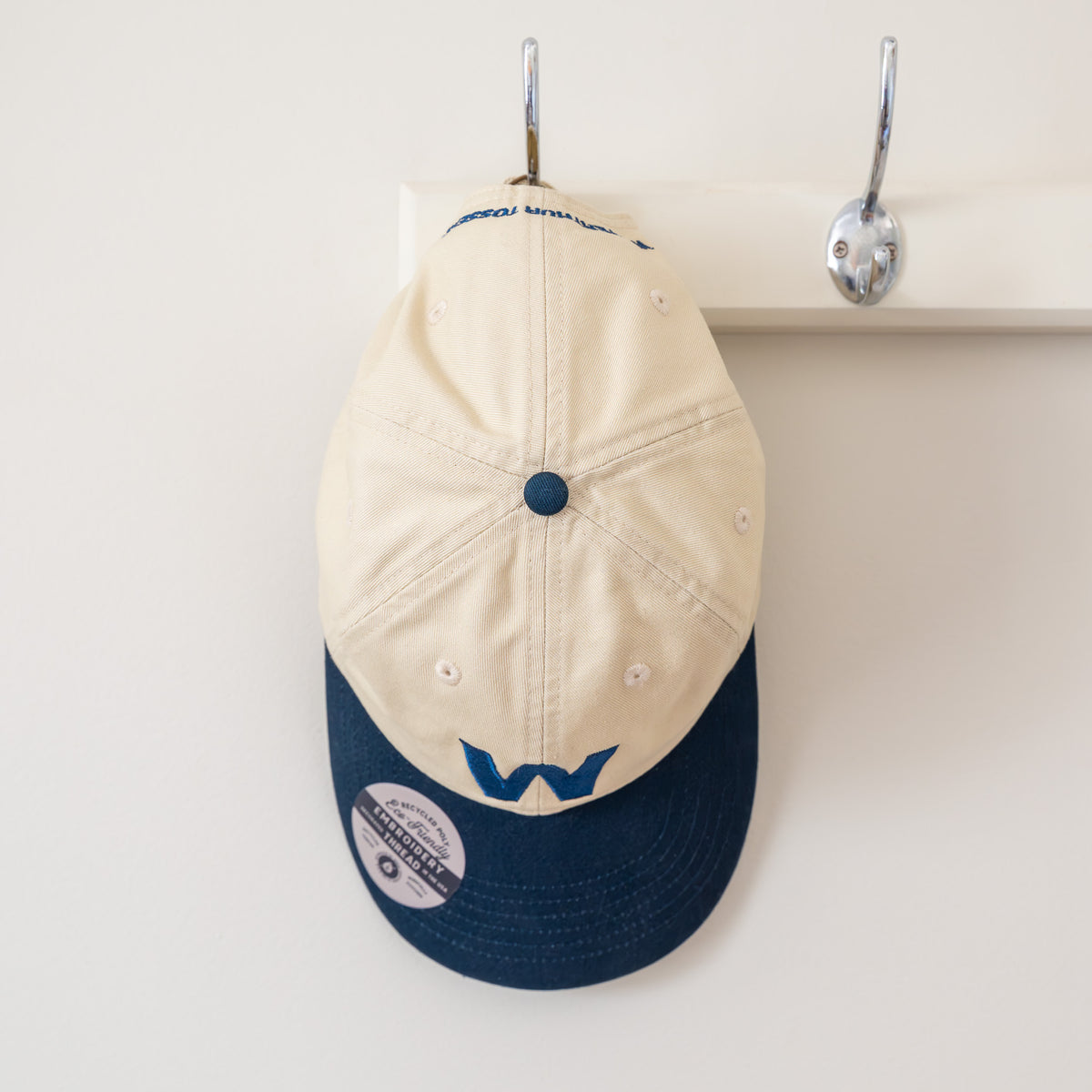 Beige and navy cap with logo on a hook against a white background