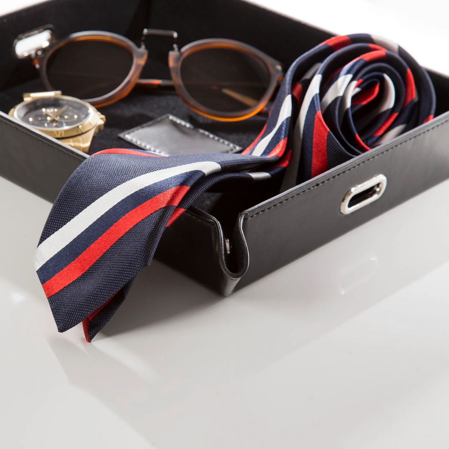 Black leather valet tray holding a striped tie, sunglasses and a wristwatch against a white background.