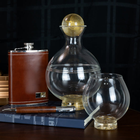 Clear glass decanter and matching drinking glass with gold accenting sitting atop a navy blue book with a wooden pen and brown leather flask with a dark background