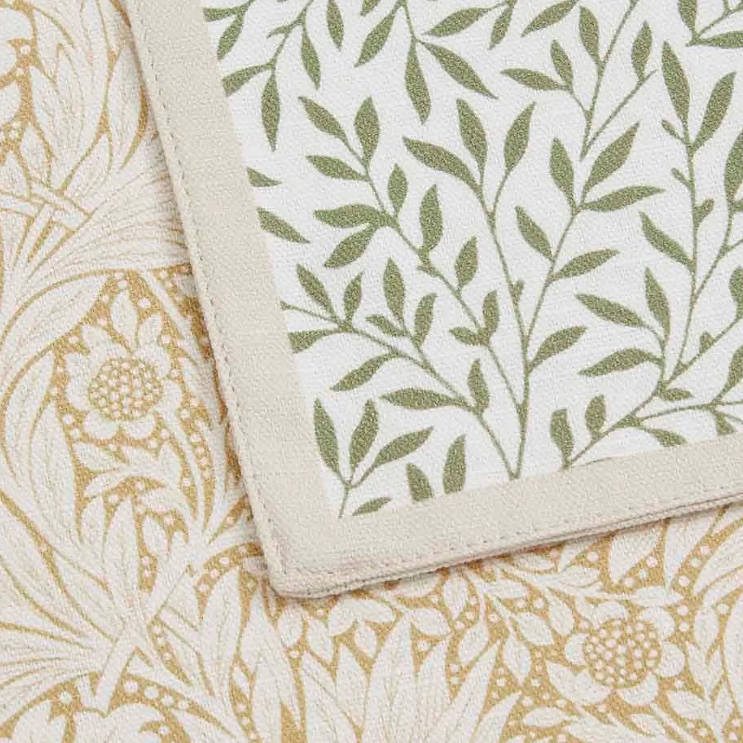 Closeup detail of two napkin, one featuring a dense, repeating botanical pattern of small green leaves and vines on a light off-white background with a light beige outer border, and the other featuring a more intricate, larger-scale floral and foliage pattern in muted yellow tone, finished with a solid muted yellow border.