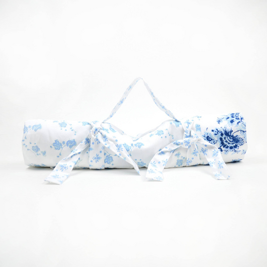 White and blue floral travel picnic blanket rolled up and tied with attached matching straps and carrying handle against a white background
