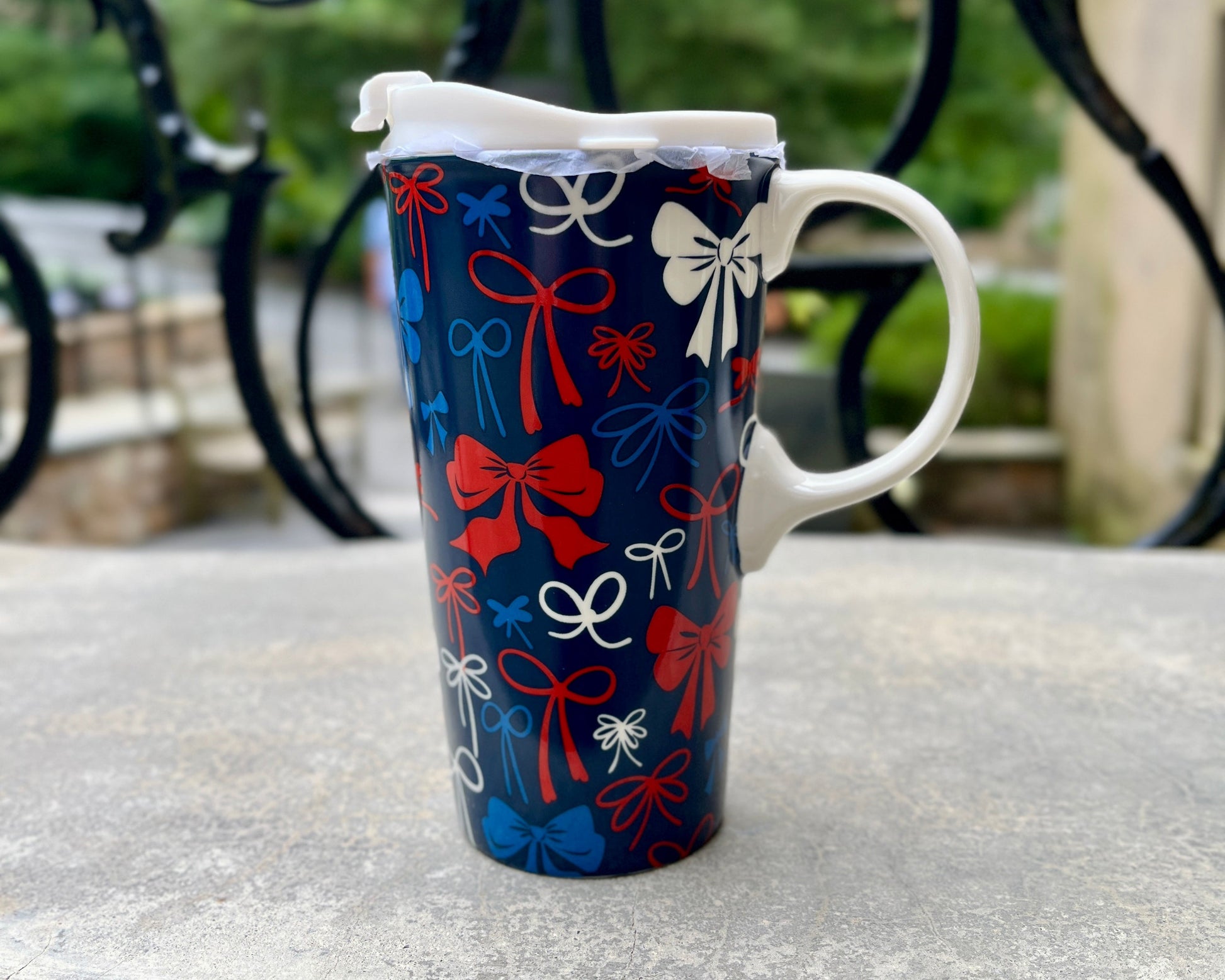 A tall ceramic mug with a festive red, white, and blue gift bow pattern, sitting on a weathered stone table with a blurred black wrought-iron fence and green foliage in the background.