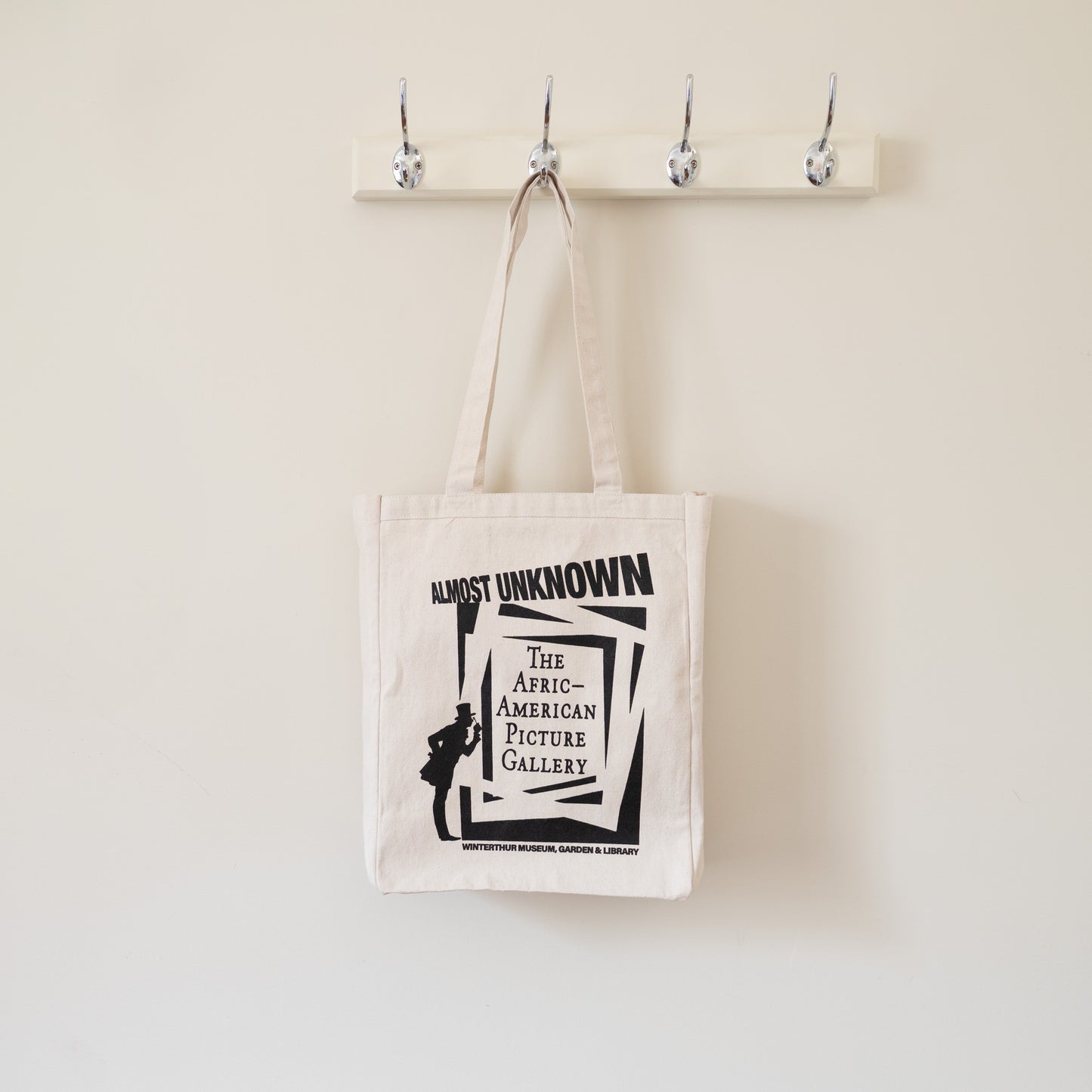 Beige tote bag with black text and graphic design hanging on a hook against a beige wall.