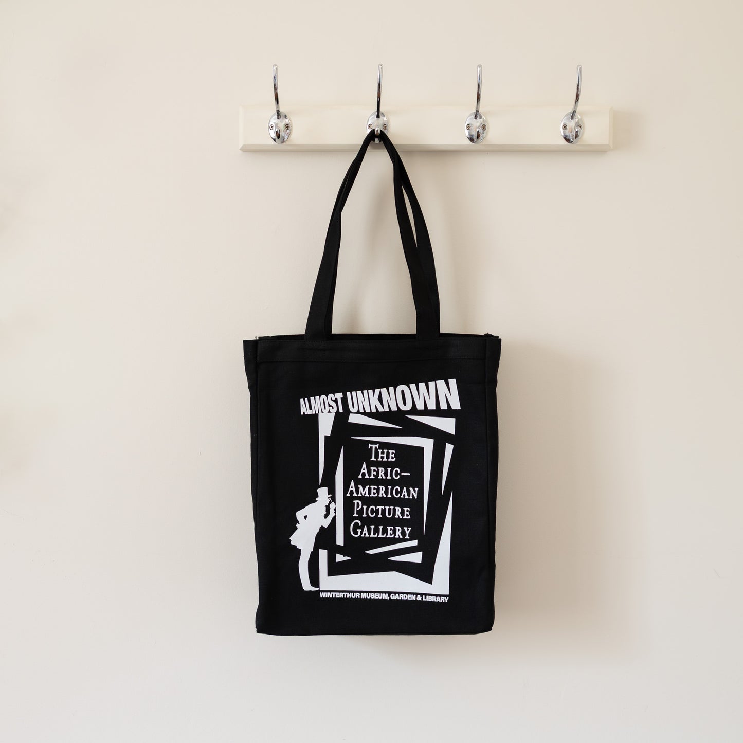 Black tote bag with white text and graphic on a beige wall with hooks.
