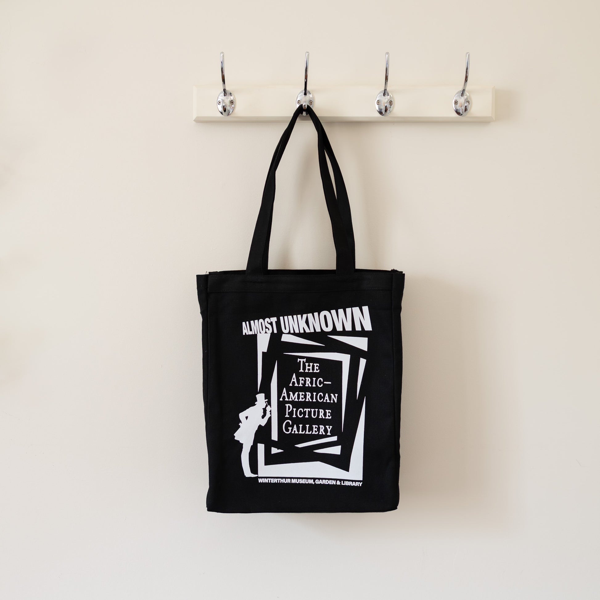 Black tote bag with white text and graphic on a beige wall with hooks.