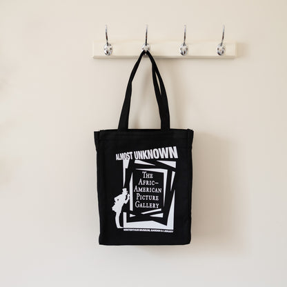 Black tote bag with white text and graphic on a beige wall with hooks.