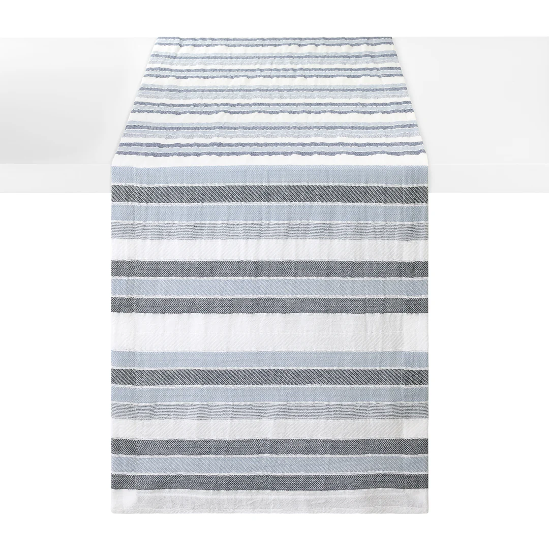 A striped woven cotton table runner with horizontal stripes in shades of blue and white, shown hanging against a white background.