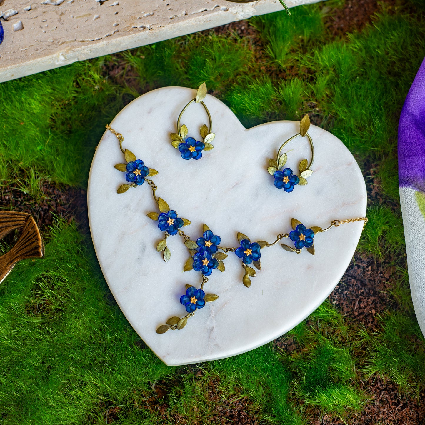 Necklace featuring blue glass flowers and green-toned sculpted bronze leaves on a gold chain on a heart-shaped marble dish atop moss.