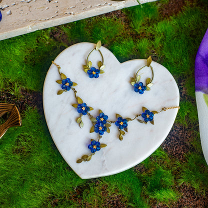 Necklace featuring blue glass flowers and green-toned sculpted bronze leaves on a gold chain on a heart-shaped marble dish atop moss.