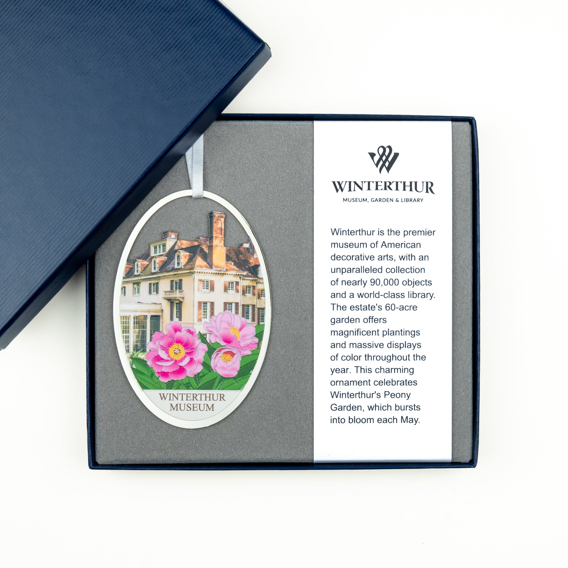 An oval ornament in a gift box from Winterthur Museum, Garden & Library, shown open on a white background with part of a navy blue box lid visible above. The ornament features a white text section with the Winterthur logo and a left-side illustration/photo of a historic mansion with pink flowers.