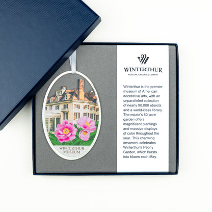 An oval ornament in a gift box from Winterthur Museum, Garden & Library, shown open on a white background with part of a navy blue box lid visible above. The ornament features a white text section with the Winterthur logo and a left-side illustration/photo of a historic mansion with pink flowers.