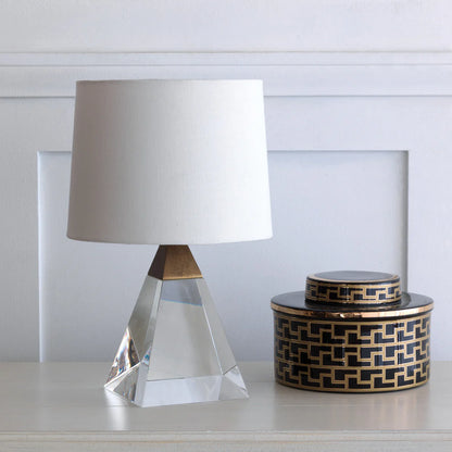 A crystal and brass pyramid-shaped lamp  with a white shade next to a round, black and gold decorative jar atop a white table