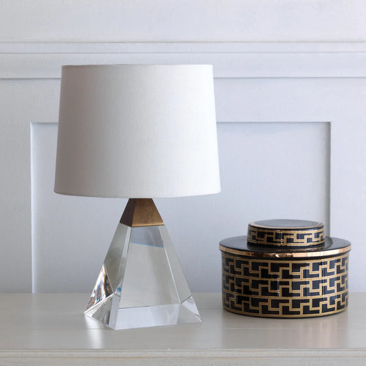 A crystal and brass pyramid-shaped lamp  with a white shade next to a round, black and gold decorative jar atop a white table
