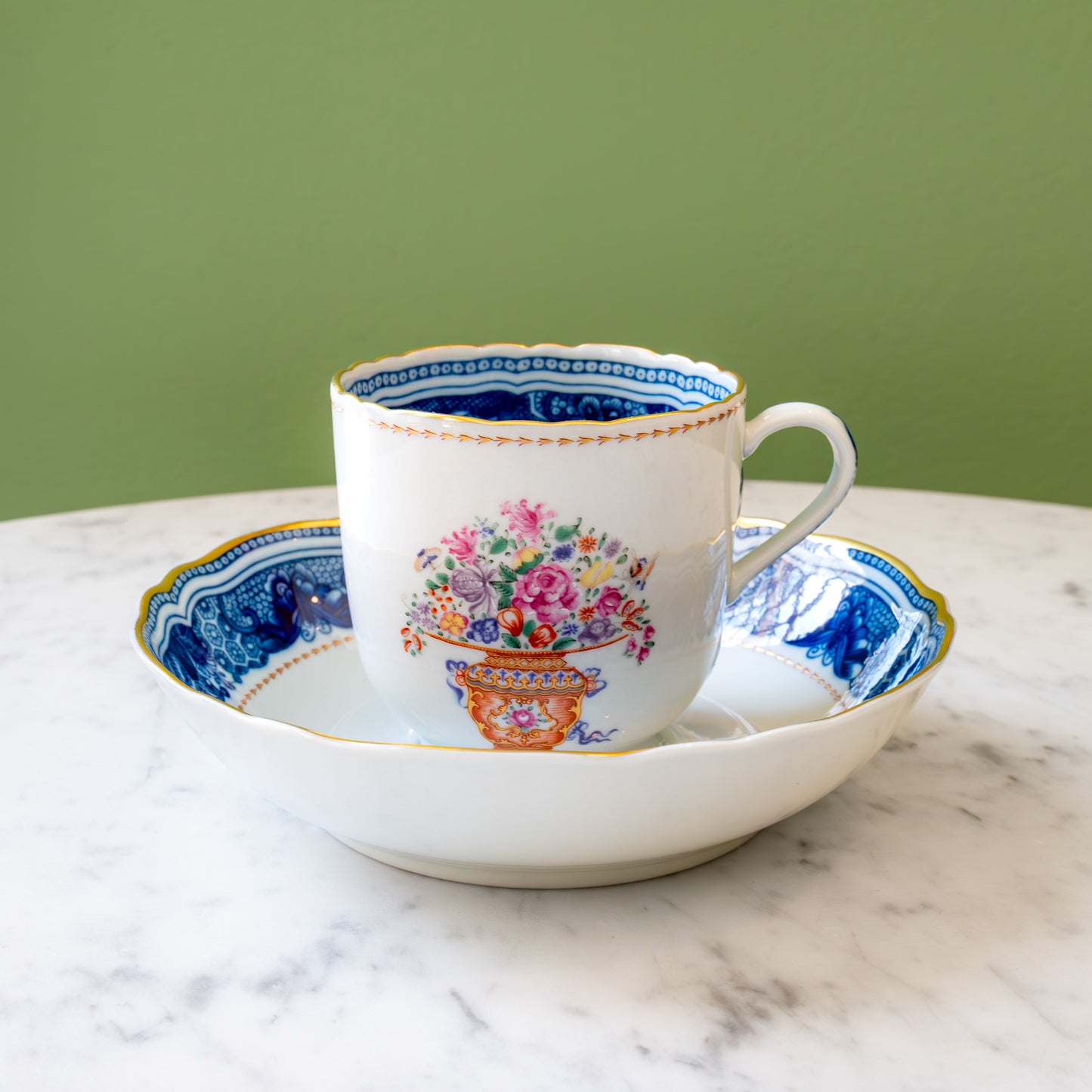 A white porcelain teacup with a colorful floral bouquet in urn motif, gold rim and decorative outer border, and blue decorative inner border sitting on a saucer with a matching blue border.