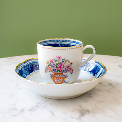 A white porcelain teacup with a colorful floral bouquet in urn motif, gold rim and decorative outer border, and blue decorative inner border sitting on a saucer with a matching blue border.