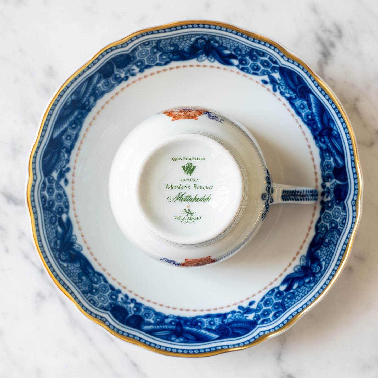 Bottom of a white ceramic teacup with branding on white saucer with a blue and gold decorative border against a marble surface.