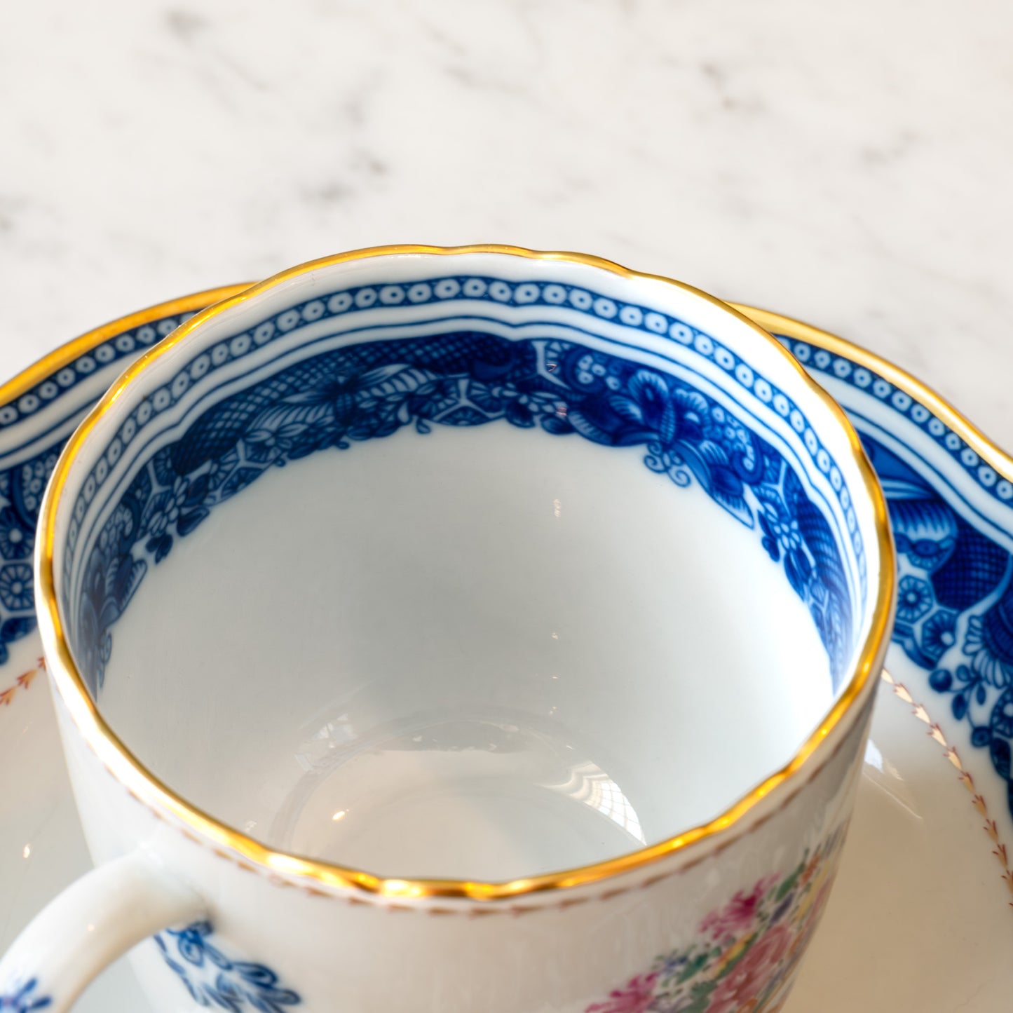 Close-up of the inner rim of a white teacup featuring decorative blue and gold patterns on a marble surface.