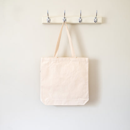 Beige tote bag hanging on a wooden hook rack against a white wall.