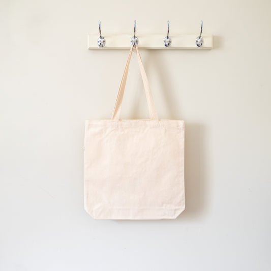 Beige tote bag hanging on a wooden hook rack against a white wall.