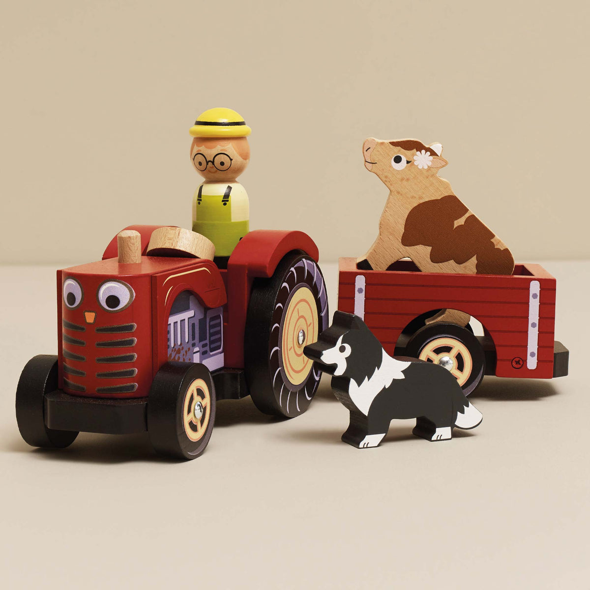 Wooden toy tractor with a driver figure, trailer with a cow, and a black and white dog on a beige background