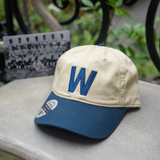 Beige and blue cap with a 'W' logo on a concrete surface outdoors with a vintage baseball team photo in the background