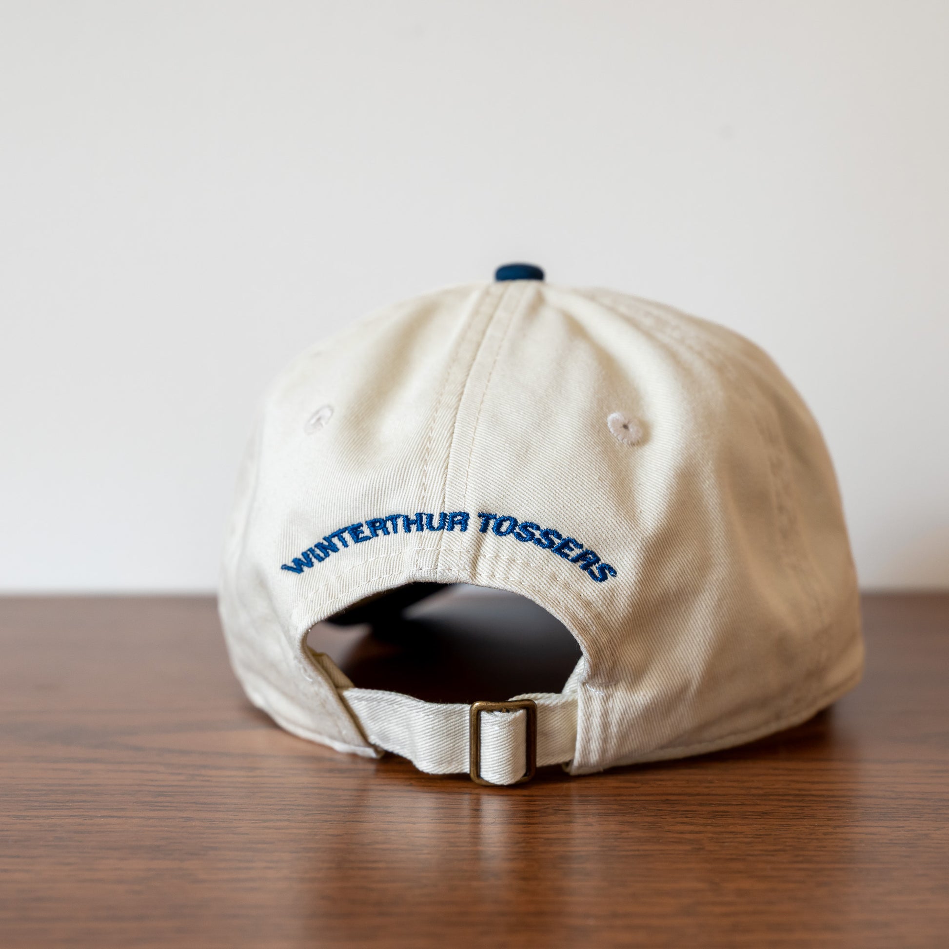 Beige cap with 'Winterthur Tossers' text  arching across the back on a wooden surface