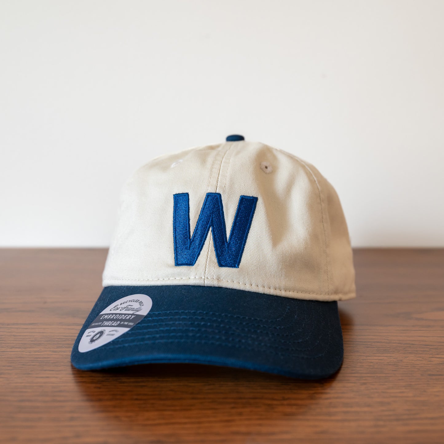 Beige cap with a blue rime and large 'W'  on the front sitting on a wooden surface