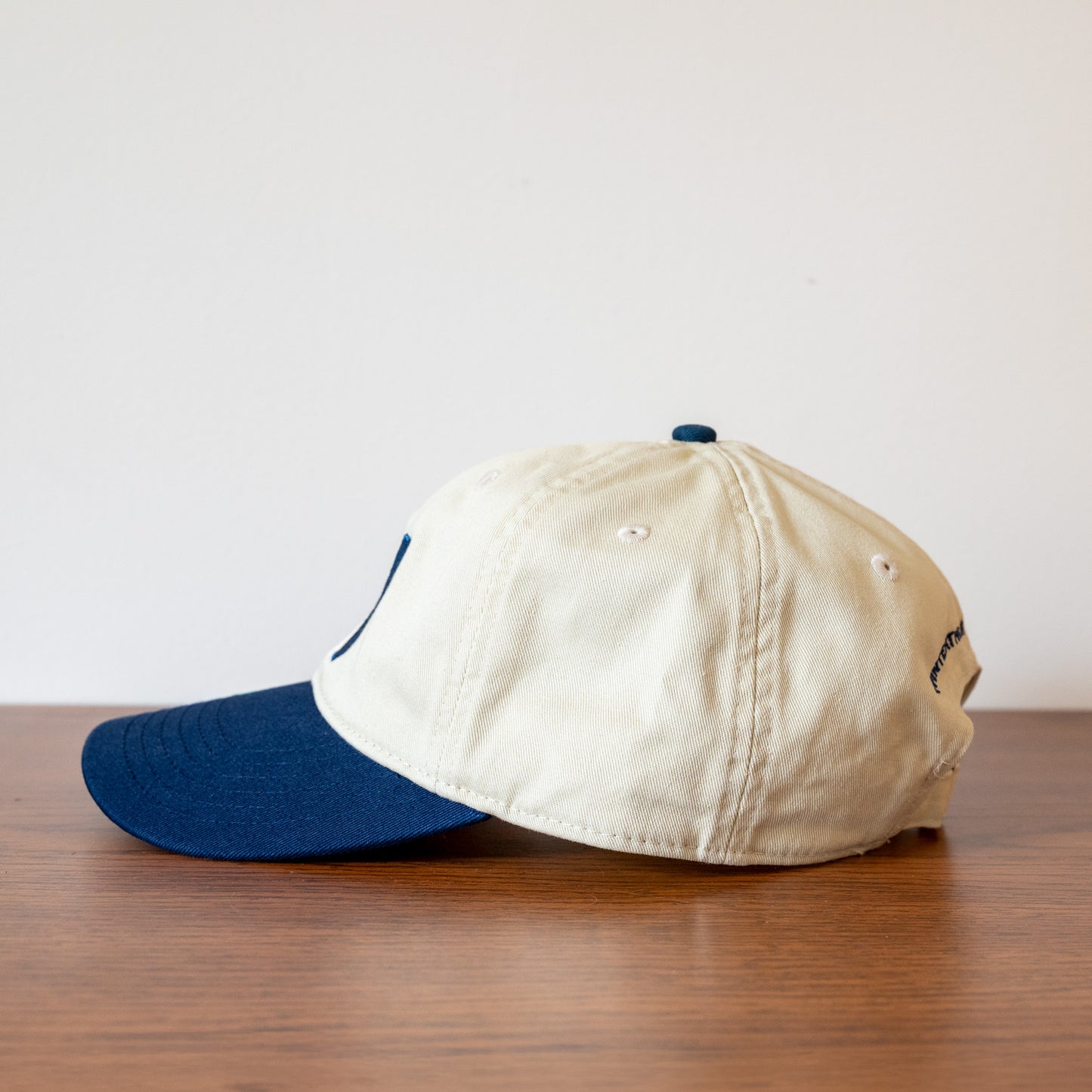 Left side of a beige and blue baseball cap on a wooden surface with a white background