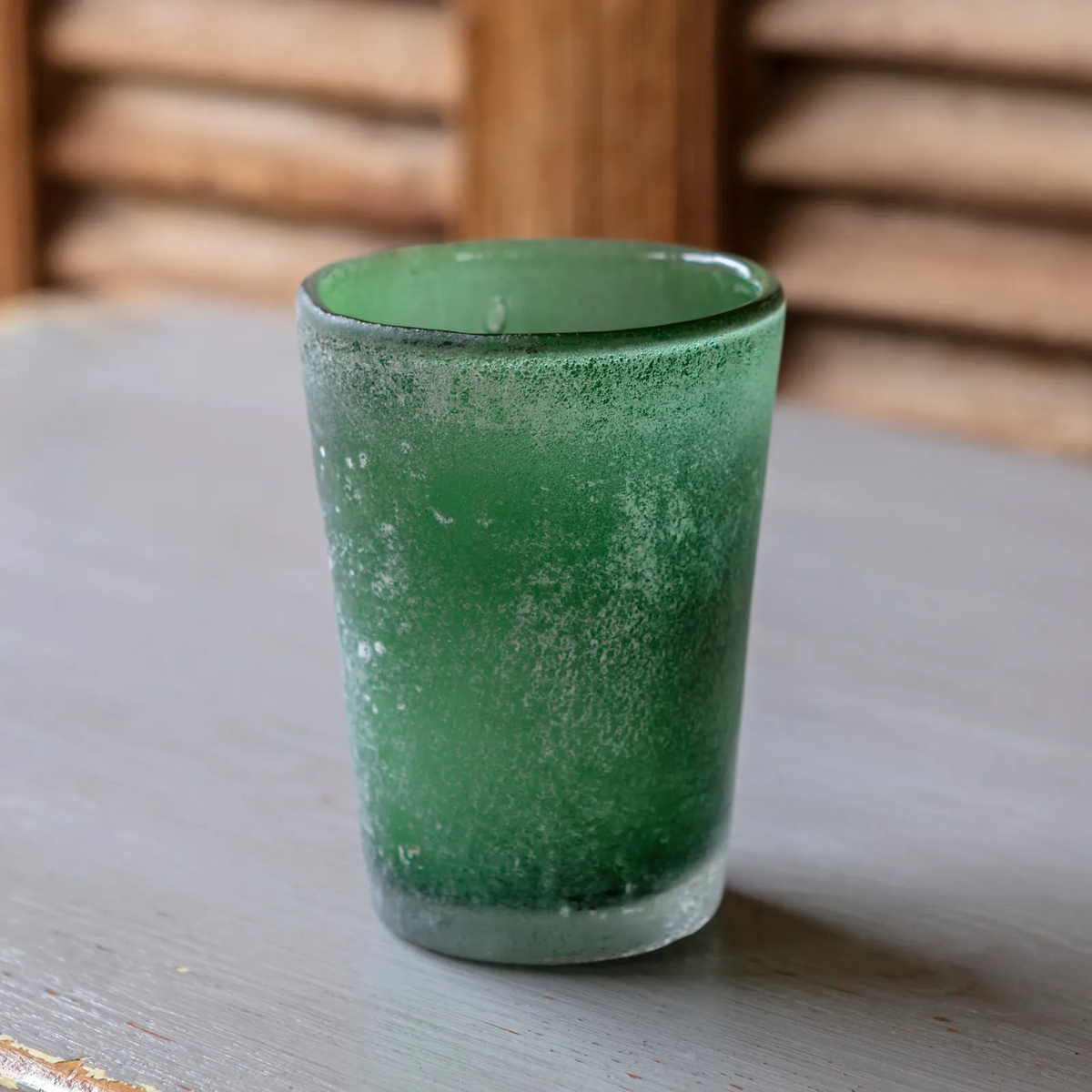 A small frosted green glass votive candle holder with a textured finish, sitting on a gray wooden table with a rustic wooden background.