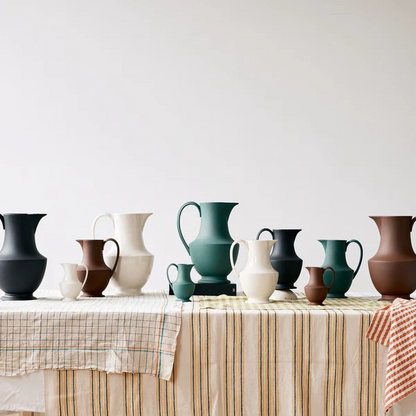 A collection of matte ceramic pitchers and vases in various colors including teal, brown, cream, and black, arranged on a table covered with striped and checkered tablecloths against a plain white wall.