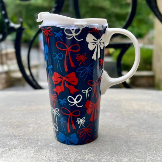 A tall ceramic mug with a festive red, white, and blue gift bow pattern, sitting on a weathered stone table with a blurred black wrought-iron fence and green foliage in the background.