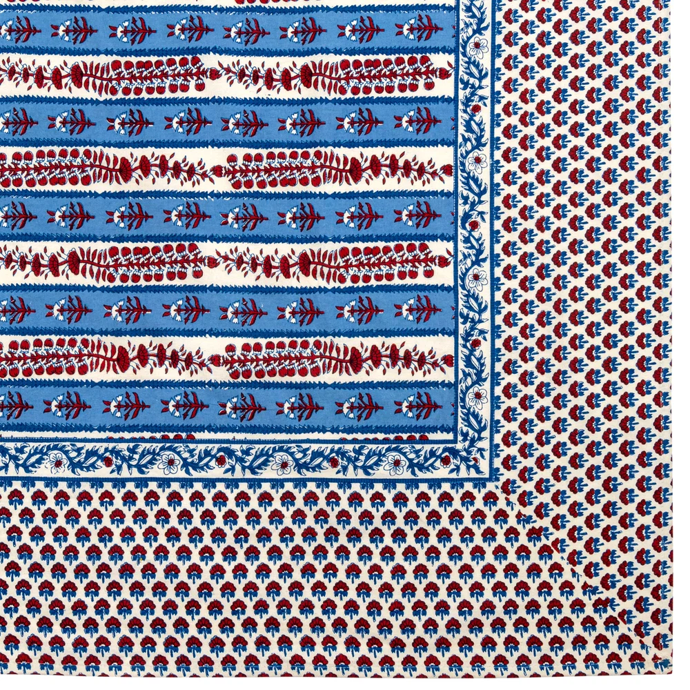 A close-up of the corner of a block printed tablecloth with repeating blue and white stripes decorated with red block-printed floral motifs and a wide white border with blue & red flowers.