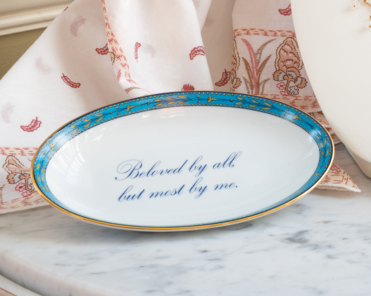 An oval porcelain tray with a blue and yellow decorative border, gold rim, and the text "Beloved by all but most by me" in blue cursive script in the center.
