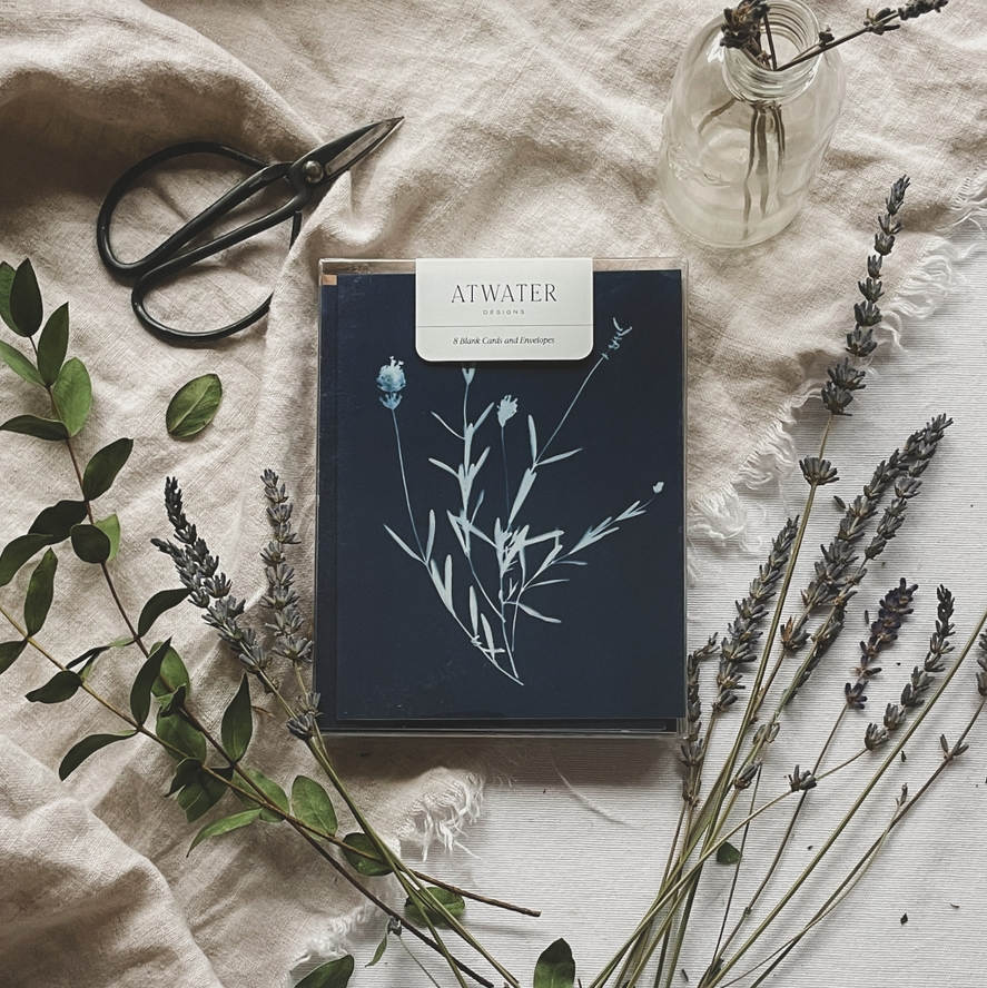 Blue and white botanical notecard set packaged in a clear box and surrounded by plants and garden sheers on a linen cloth