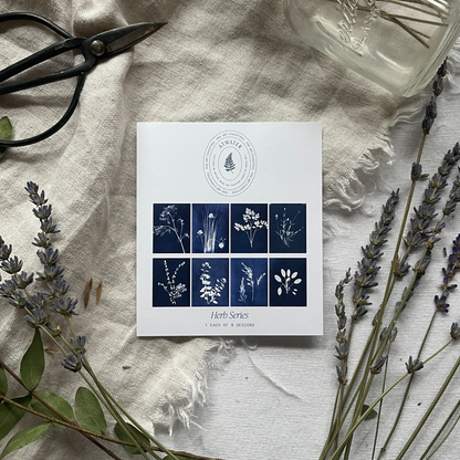 White card displaying 8 different blue and white botanical prints entitled 'Herb Series' on a linen cloth surrounded by plants and a pair of garden sheers
