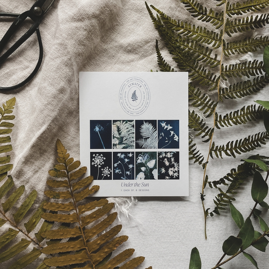 White card displaying 8 different blue and white fern prints entitled 'Under the Sun' on a linen cloth surrounded by plants and a pair of garden sheers