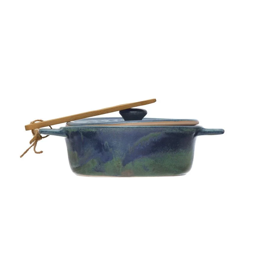 Side view of a reactive glazed blue and green lidded stoneware baking dish with a wooden spoon tied to one handle with twine against a white background