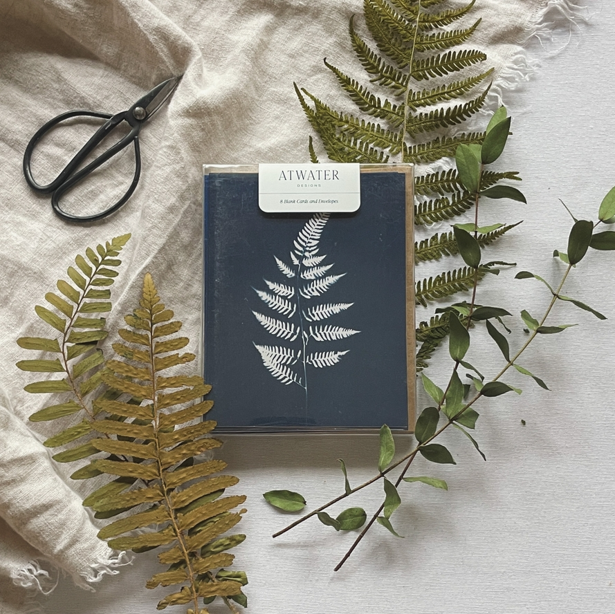 Blue and white fern printed notecard set packaged in a clear box and surrounded by plants and garden sheers on a linen cloth