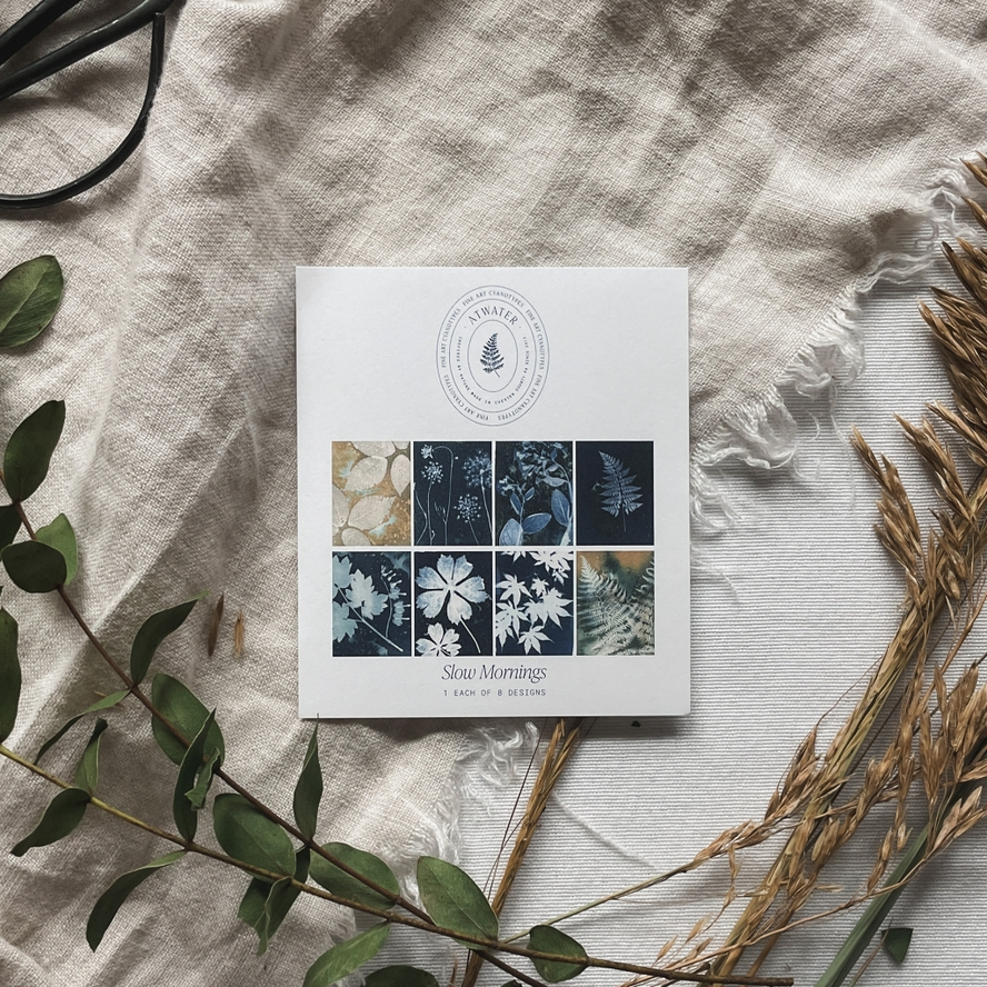 White card displaying 8 botanical cyanotype prints entitled 'Slow Mornings' on a linen cloth surrounded by plants