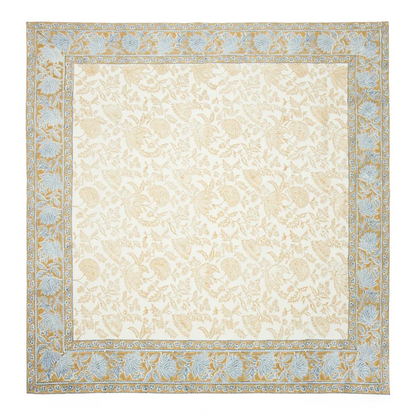 A cream-colored square tablecloth featuring a central block-printed floral pattern in a muted deep mustard yellow framed with a deep mustard border with a repeating blue floral print.