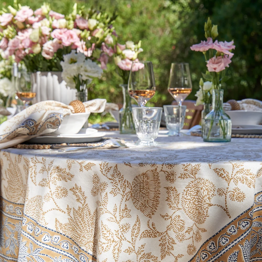 A cream-colored tablecloth featuring a block-printed floral pattern in a muted deep mustard and blue shown draped over a table outdoors and decorated with dinnerware and floral arrangements.