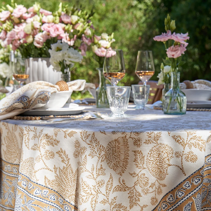 A cream-colored tablecloth featuring a block-printed floral pattern in a muted deep mustard and blue shown draped over a table outdoors and decorated with dinnerware and floral arrangements.