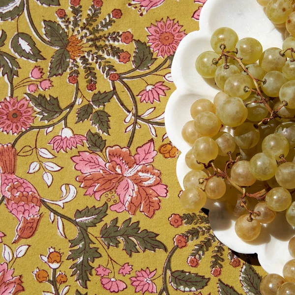 Close-up of a mustard-colored tablecloth with a traditional floral block-printed pattern in olive green, saffron, and rose pink with a white plate holding green grapes sitting on top.