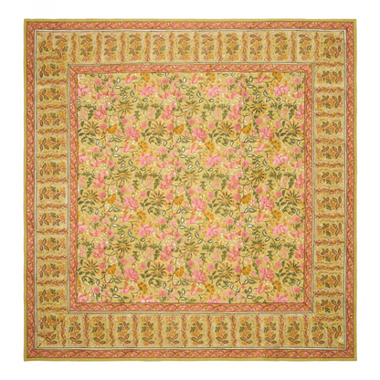 A square mustard-colored tablecloth with a traditional block-printed floral pattern in olive green, saffron, and rose pink, and a contrasting repeated leaf border laid flat against a white background.