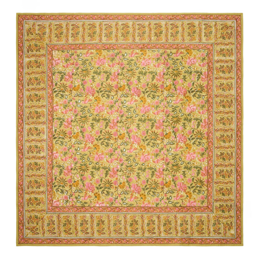 A square mustard-colored tablecloth with a traditional block-printed floral pattern in olive green, saffron, and rose pink, and a contrasting repeated leaf border laid flat against a white background.