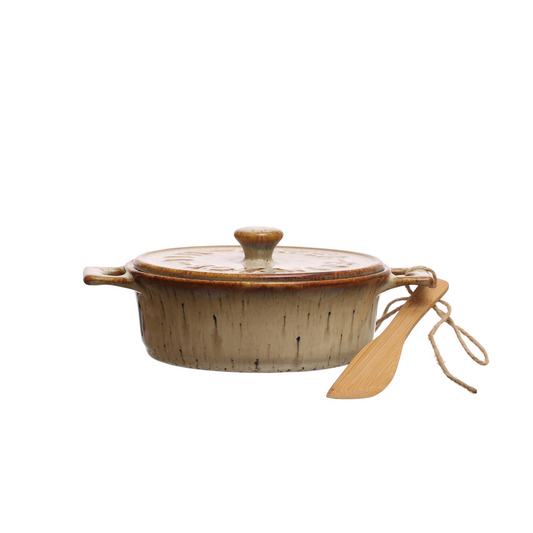 Side view of a brown speckled stoneware lidded baking dish with a wooden spoon tied to one handle with twine against a white background