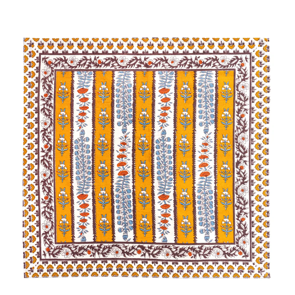 Square cloth napkin with a mustard yellow, off-white and mahogany brown striped pattern and decorated with block printed light blue and burnt orange floral motifs laid flat against a white background