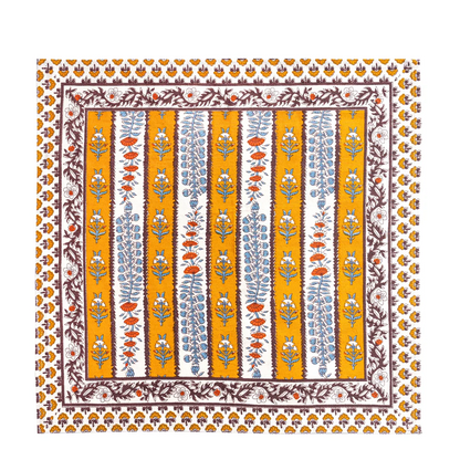 Square cloth napkin with a mustard yellow, off-white and mahogany brown striped pattern and decorated with block printed light blue and burnt orange floral motifs laid flat against a white background