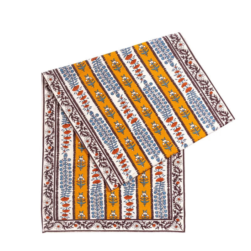 Folded table runner featuring an off-white and mustard yellow striped pattern decorated with mahogany brown, light blue and burnt orange floral block printed motifs against a white background.