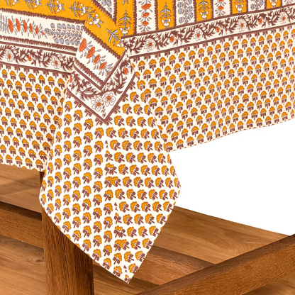 A close-up of a printed tablecloth draped over a wooden table, showing repeating yellow, off-white and brown stripes decorated with block-printed floral motifs in light blue and orange.
